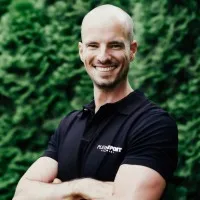 Smiling man with crossed arms in a black polo, standing before a leafy green background.