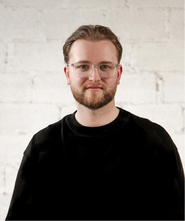 Portrait of a bearded man in glasses, black top, standing against a light brick wall.