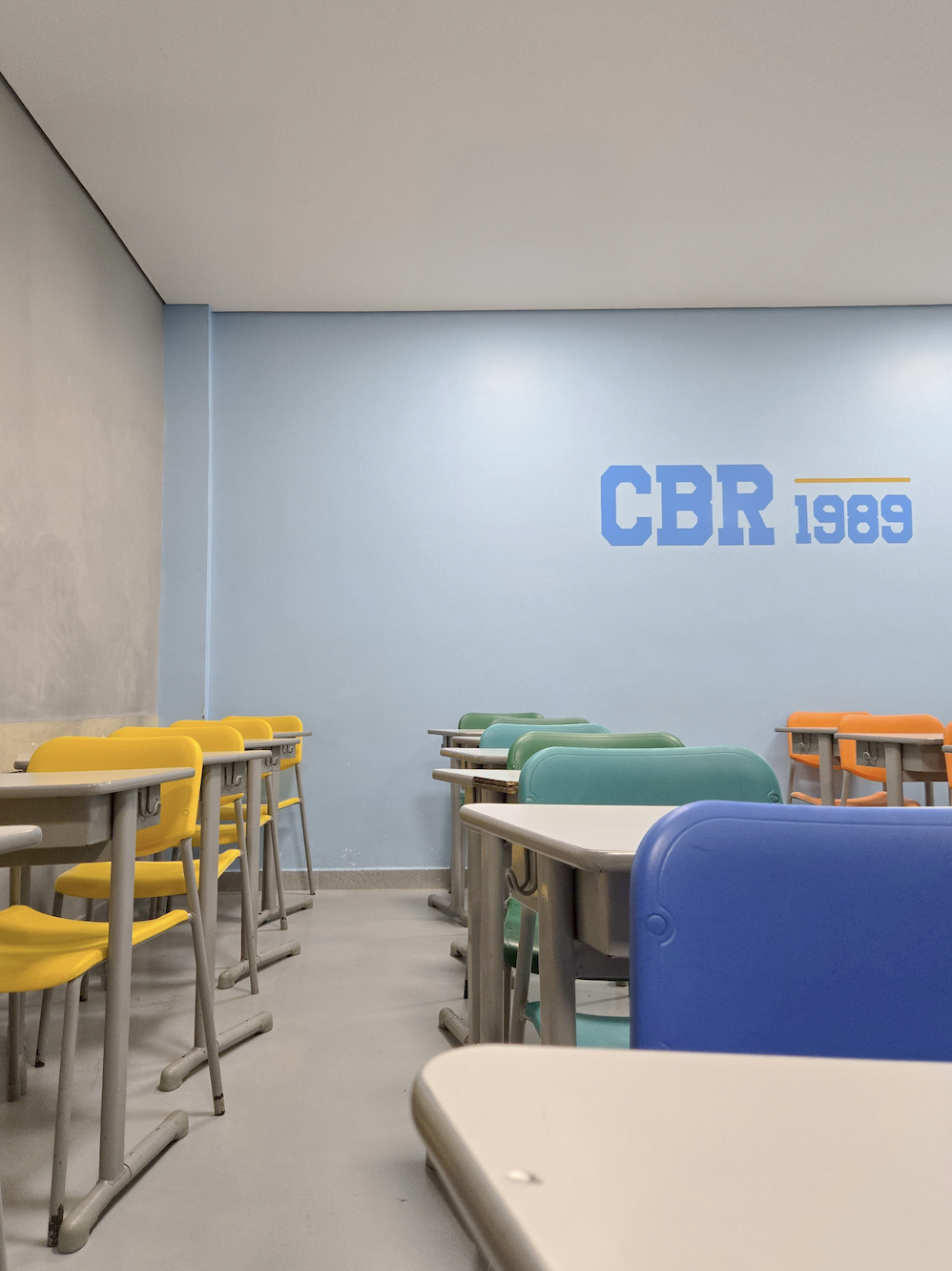 Sala de aula moderna e colorida do Colégio Bom Retiro (CBR), com carteiras escolares em tons vibrantes de amarelo, verde, laranja e azul, promovendo um ambiente estimulante e acolhedor. A parede azul clara traz o logo “CBR 1989”, reforçando a identidade da escola. O espaço é bem iluminado, com piso cinza e layout organizado, ideal para incentivar o aprendizado com conforto e criatividade.
