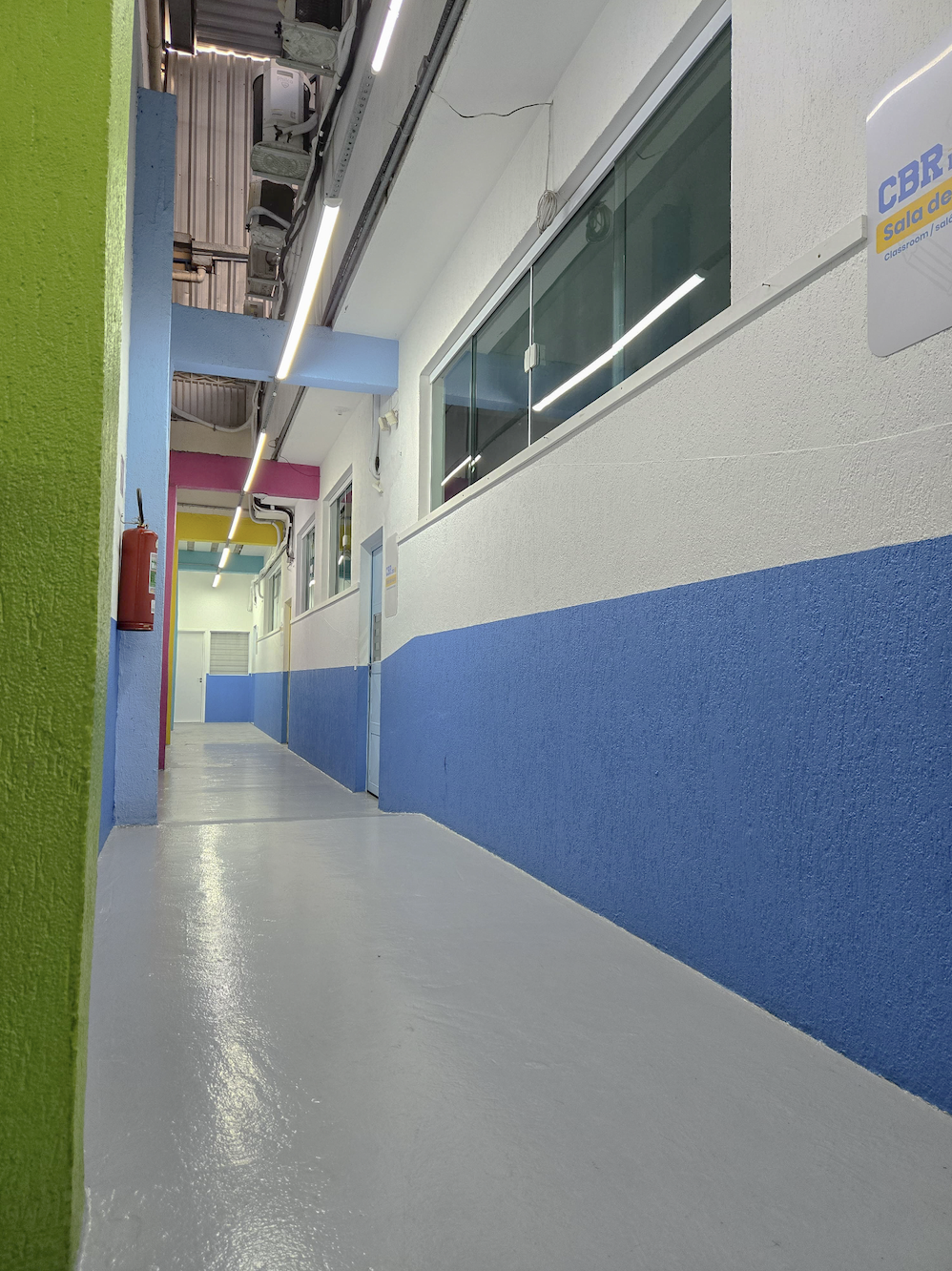 Corredor interno de escola com pintura colorida e moderna nas paredes, combinando tons de azul, verde e detalhes em rosa e amarelo. O espaço é limpo, bem iluminado com luzes LED no teto e apresenta janelas de vidro que conectam as salas ao corredor. Na parede, há uma placa identificando o ambiente como parte do CBR (Colégio Bom Retiro), reforçando organização, cuidado estético e ambiente escolar acolhedor.