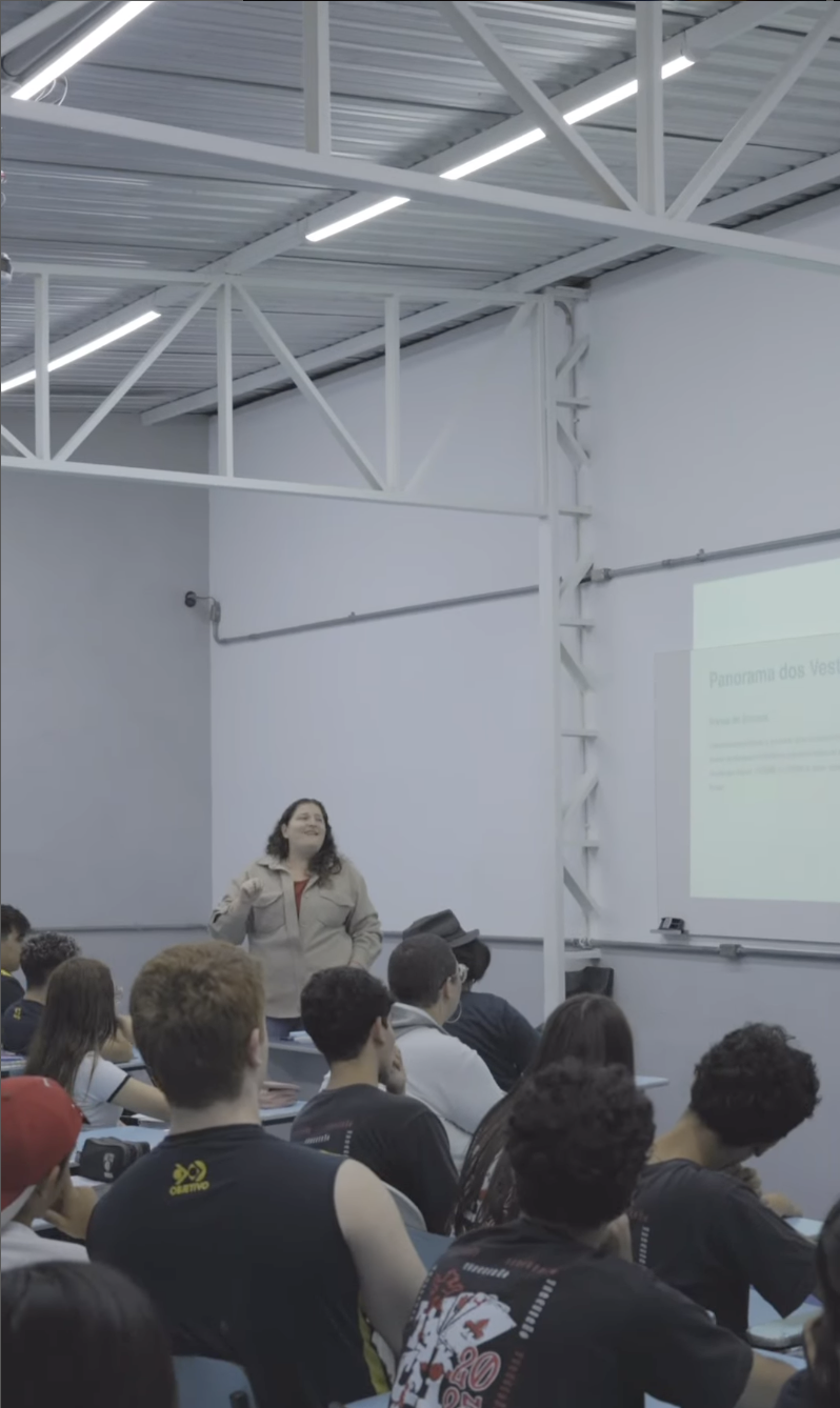 Sala de aula com estudantes do ensino médio assistindo a uma apresentação sobre “Panorama dos Vestibulares”. Professora em destaque conduz a aula de forma engajada, utilizando projetor e recursos visuais. Ambiente moderno com estrutura metálica e iluminação LED. Alunos vestem camisetas do Colégio Objetivo e uniformes escolares. Cena representa educação, preparação para vestibulares e ambiente escolar participativo.