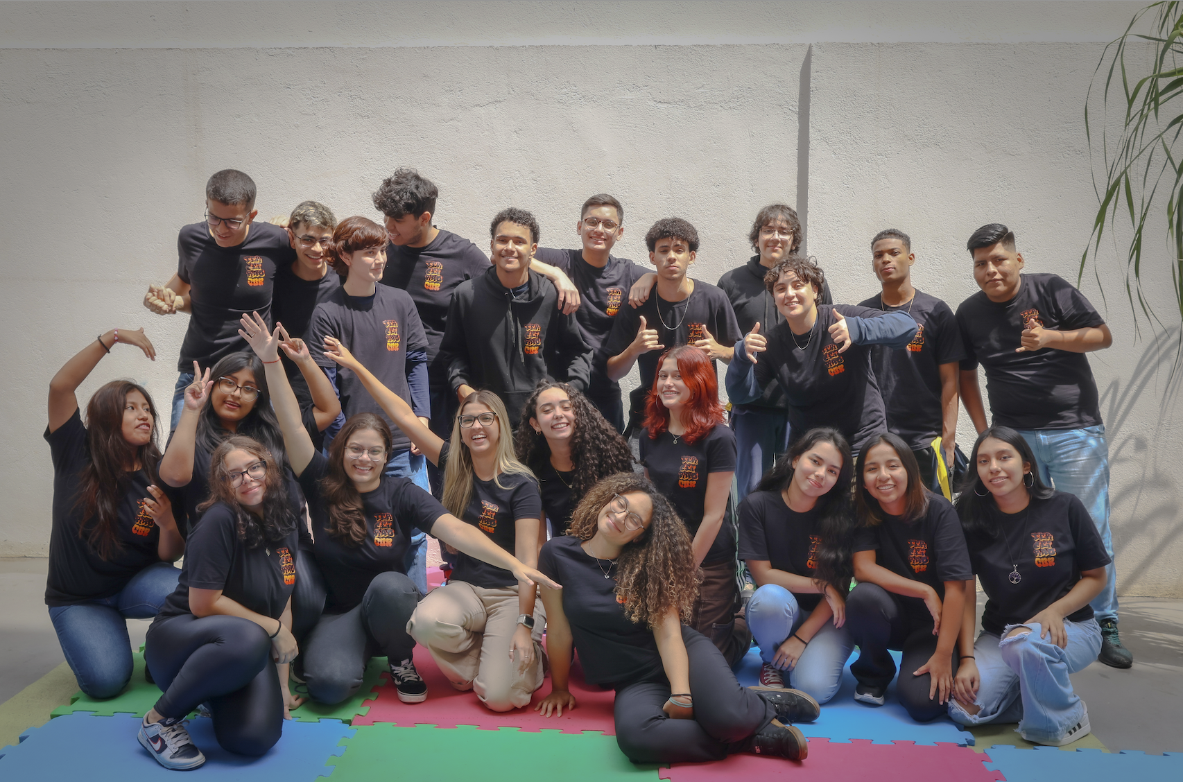 Turma de alunos do ensino médio posa de forma descontraída para foto em grupo. Jovens usam camisetas personalizadas de formandos em ambiente escolar, demonstrando união, alegria e espírito de equipe. A imagem foi tirada em espaço aberto, com piso emborrachado colorido, transmitindo energia positiva, amizade e celebração da trajetória estudantil.