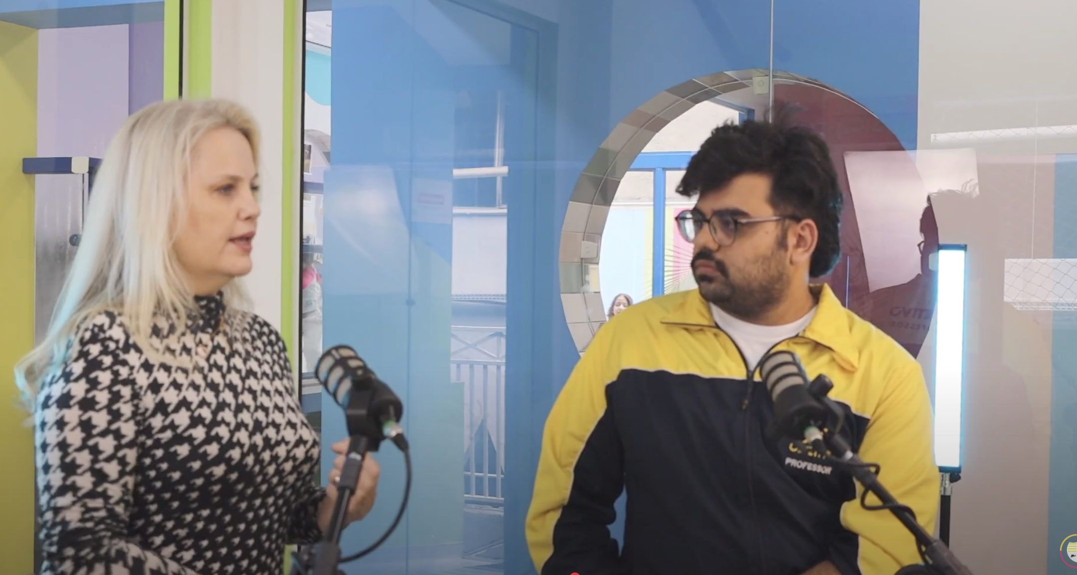 Gravação de podcast educacional realizada no estúdio do Colégio Bom Retiro (CBR). A imagem mostra dois participantes — uma mulher loira e um homem de óculos com jaqueta amarela e azul — em frente a microfones profissionais, discutindo temas pedagógicos em um ambiente moderno e colorido. O cenário tem elementos vibrantes e identidade visual institucional ao fundo, reforçando inovação, comunicação e educação interativa.
