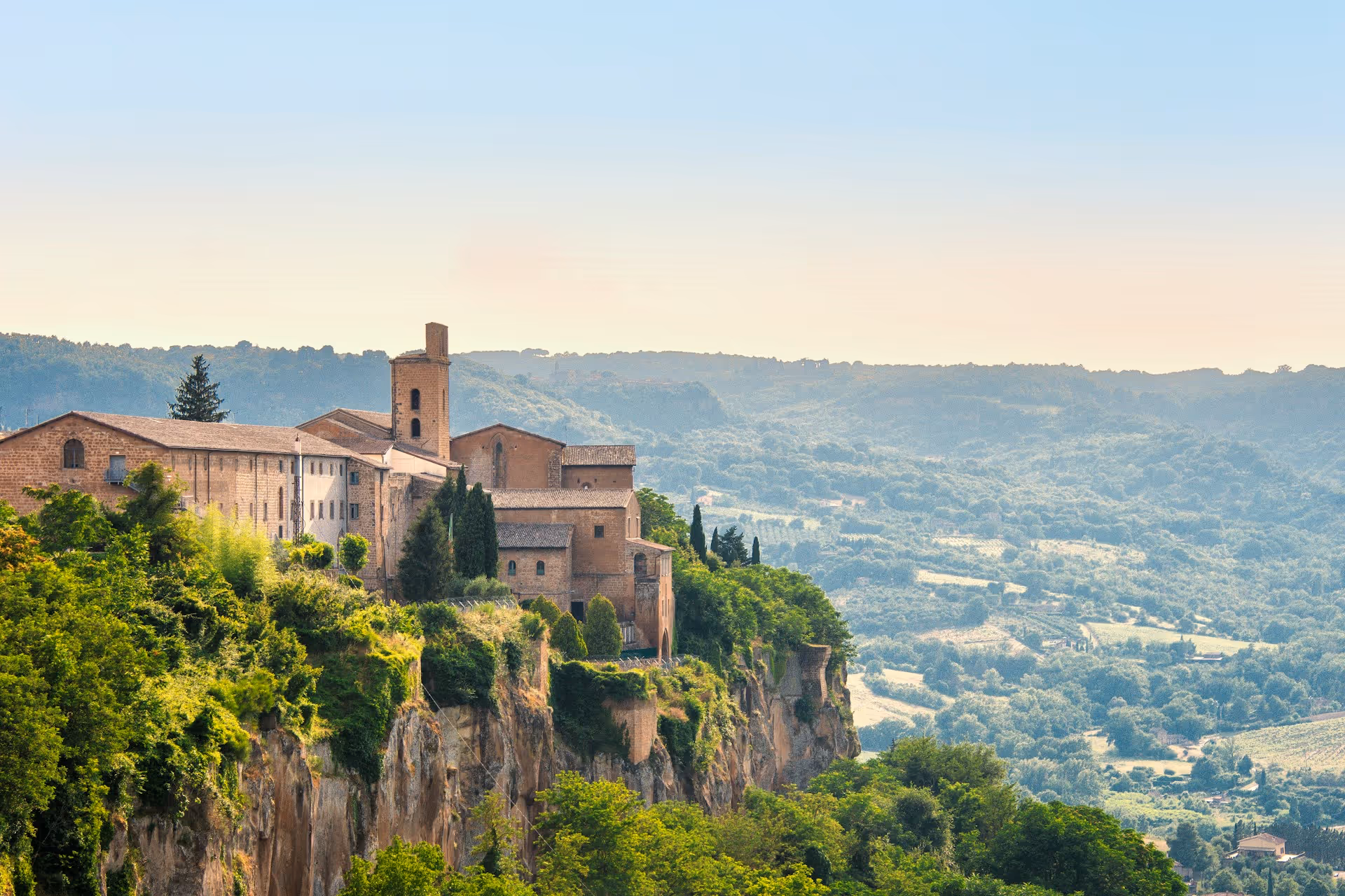Historic stone buildings perched on a green cliff overlooking a vast valley of trees and hills under a clear sky.