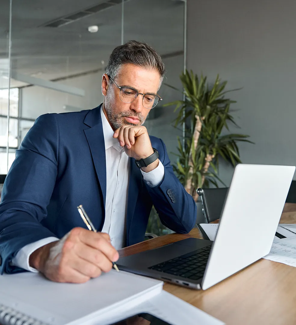 Man in a blue suit and glasses writing in a notebook at a desk with an open laptop and documents, in an office setting.