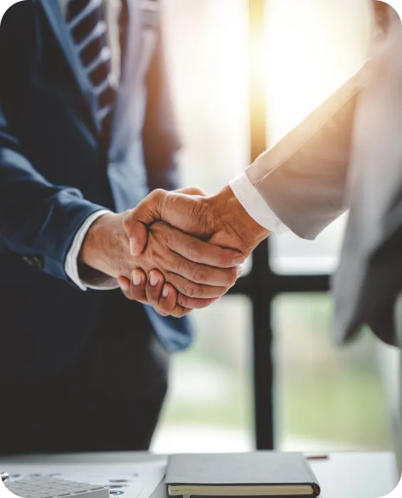 Two business people in suits shaking hands over a desk with a notebook and calculator, illuminated by sunlight from a window.