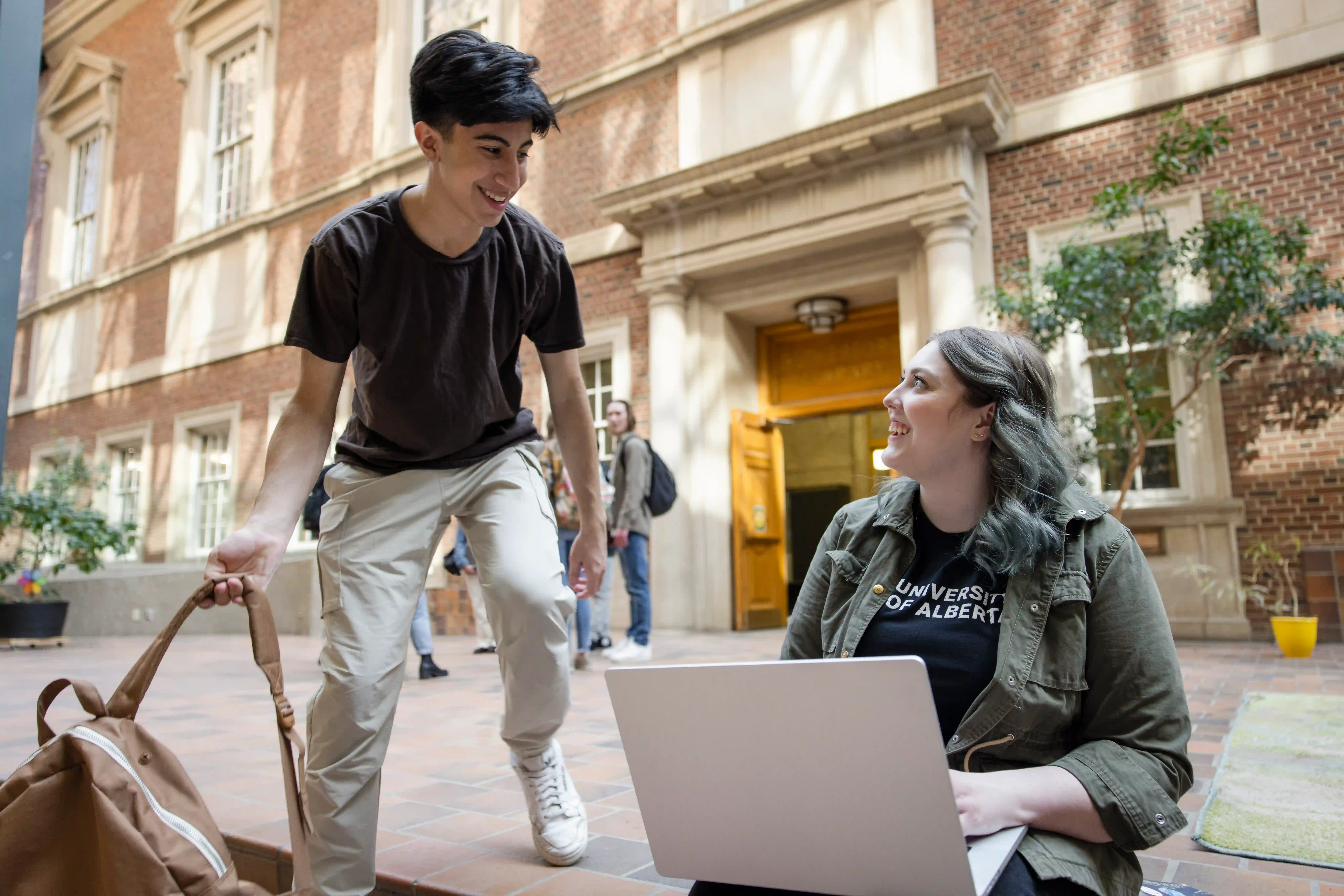 A student about to sit down with another student holding a laptop
