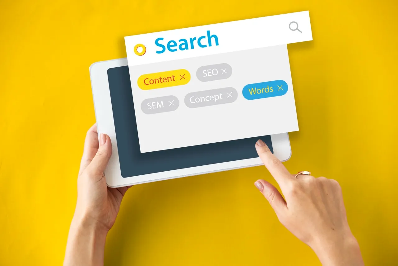 Une main tient une tablette blanche avec un écran de recherche affichant des mots-clés SEO sur fond jaune.