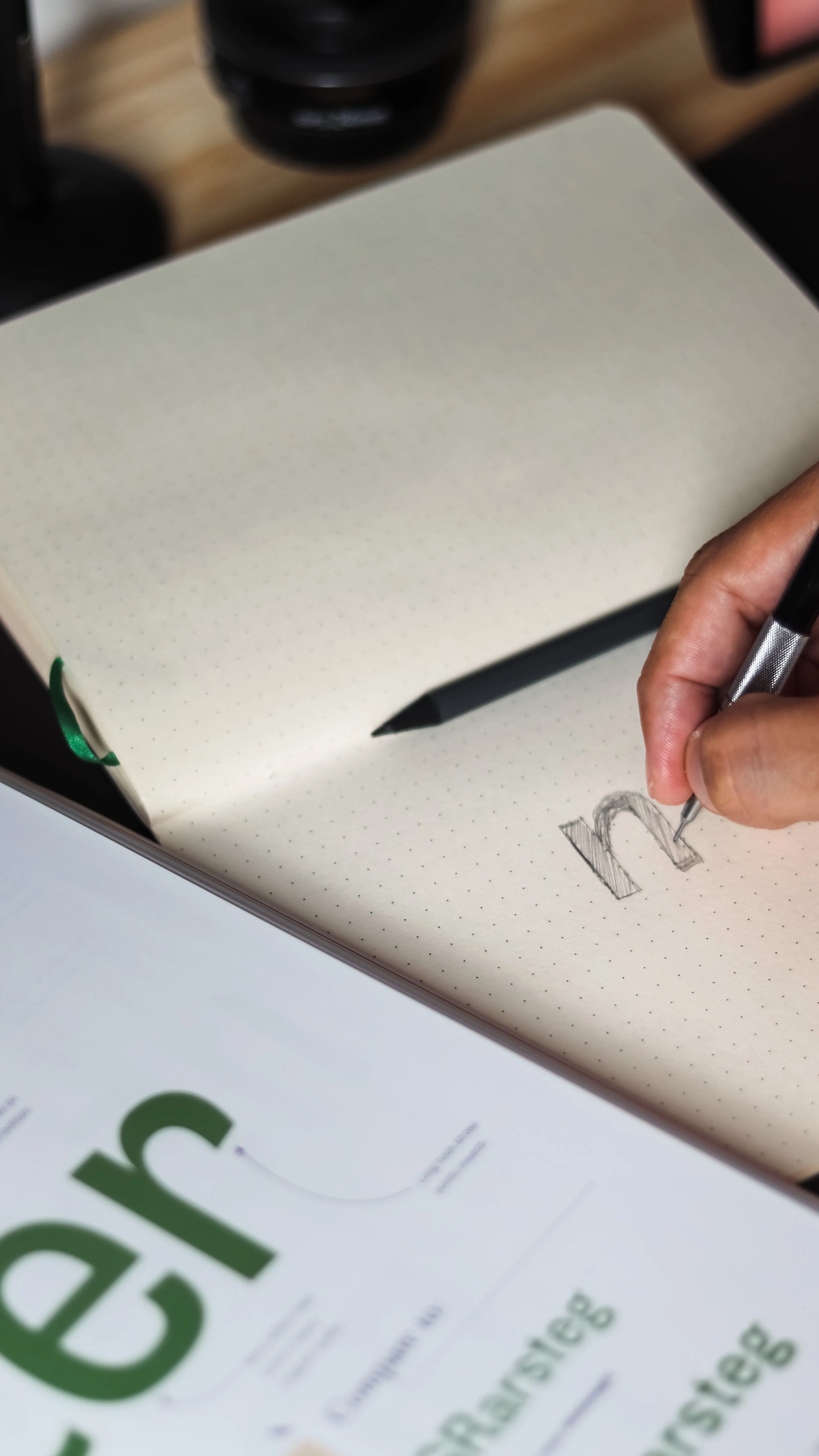 Main gauche tenant un stylo dessinant la lettre m en majuscule dans un carnet à points.