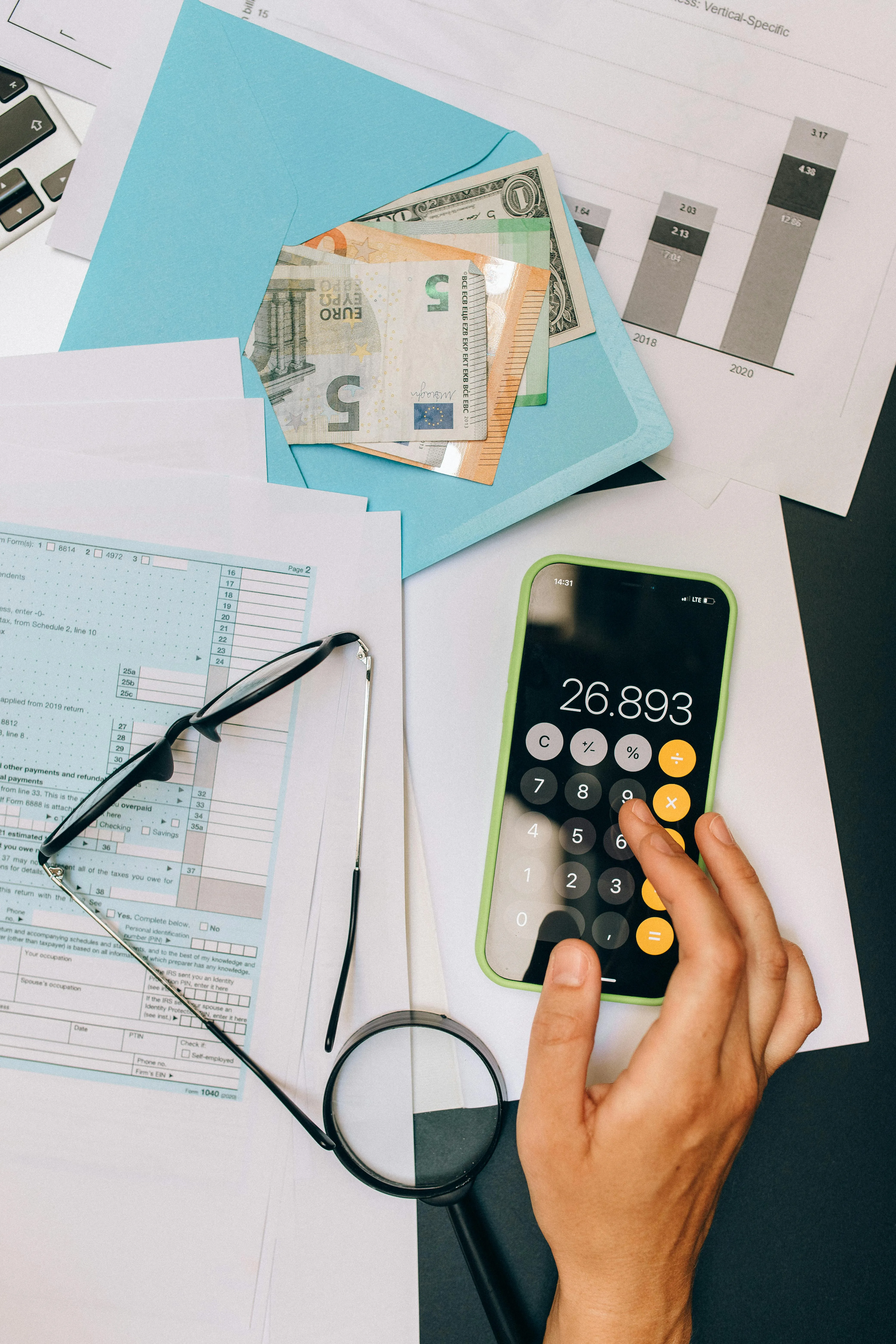 Main tenant une calculatrice sur un smartphone, entouré de documents, lunettes, argent et graphiques.