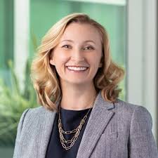 Professional headshot of blonde woman in gray blazer and gold necklace smiling against green office background