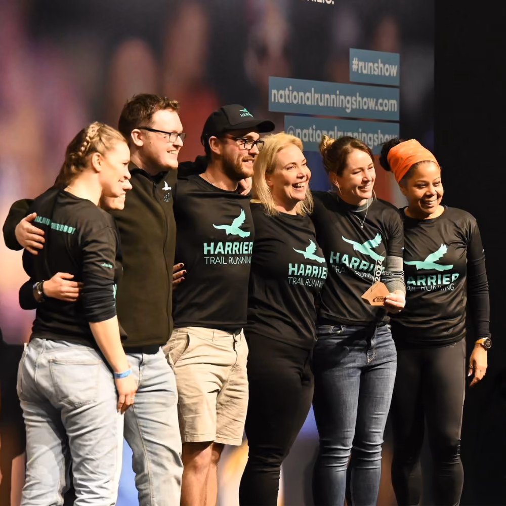 Group of six smiling runners posing on stage wearing Harrier Trail Running shirts holding a wooden award.