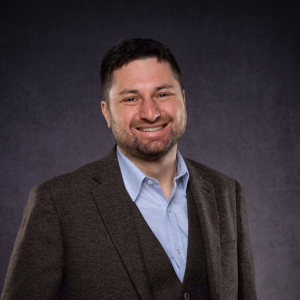Smiling man with short hair and beard wearing a brown suit jacket and light blue shirt against gray background.