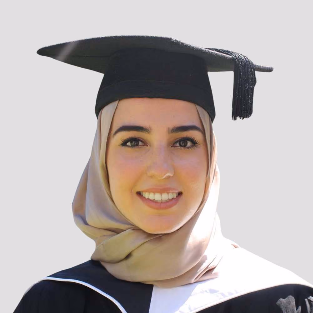 Young woman wearing a beige hijab and black graduation cap and gown, smiling on gray background.