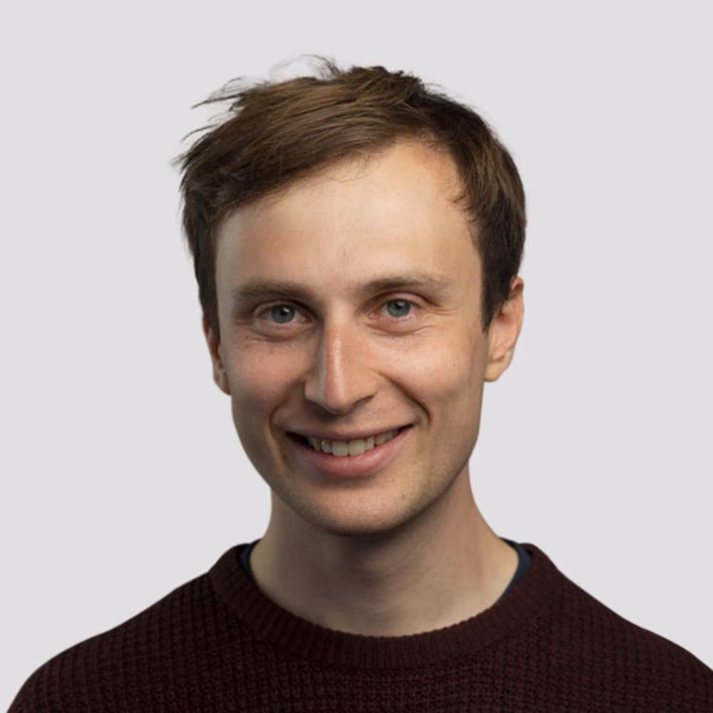 Smiling young man with light skin, short brown hair, and blue eyes wearing a dark sweater.