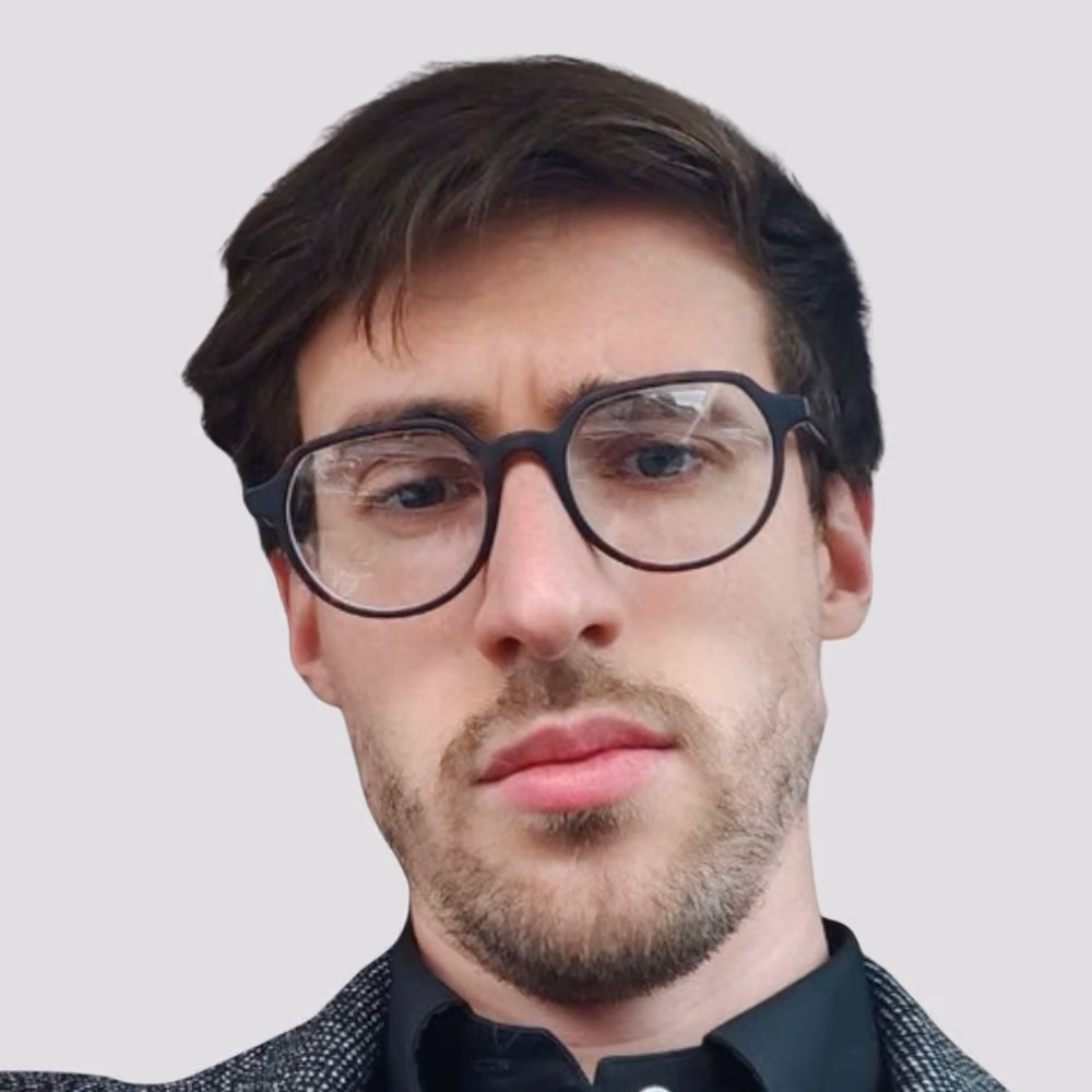 Close-up of a man with glasses, short hair, and light facial hair against a plain background.