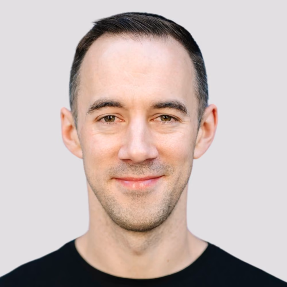Smiling man with short dark hair and light beard, wearing a black shirt on a gray background.