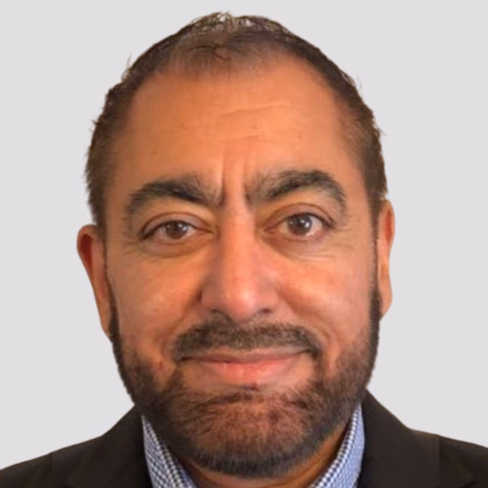 Close-up of a smiling man with a beard wearing a checked shirt and dark jacket against a plain background.