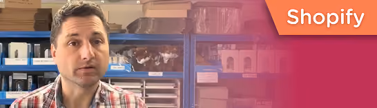 Man in a plaid shirt speaking in a room with shelves and packing supplies, Shopify logo in corner.