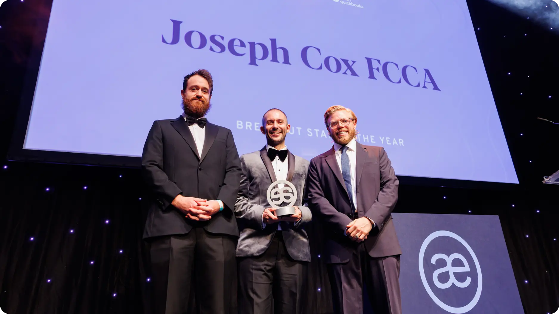 Three men in suits stand on stage smiling, one holding an award under a screen with text.