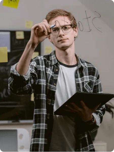 Un jeune homme portant des lunettes et une chemise à carreaux écrit sur une surface en verre tout en tenant une tablette.