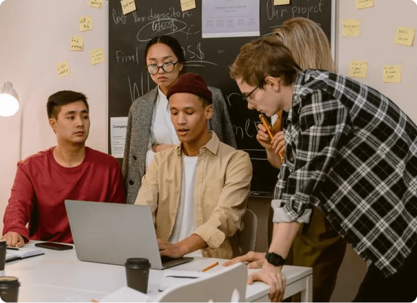 Groupe de cinq jeunes collaborateurs travaillant ensemble autour d'un ordinateur portable dans un bureau, avec un tableau noir et des post-it en arrière-plan.