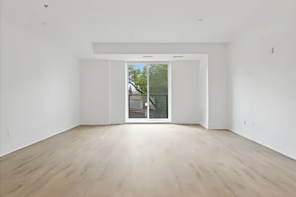 Grand salon vide avec sol en bois clair, murs blancs et grande baie vitrée donnant sur des arbres.