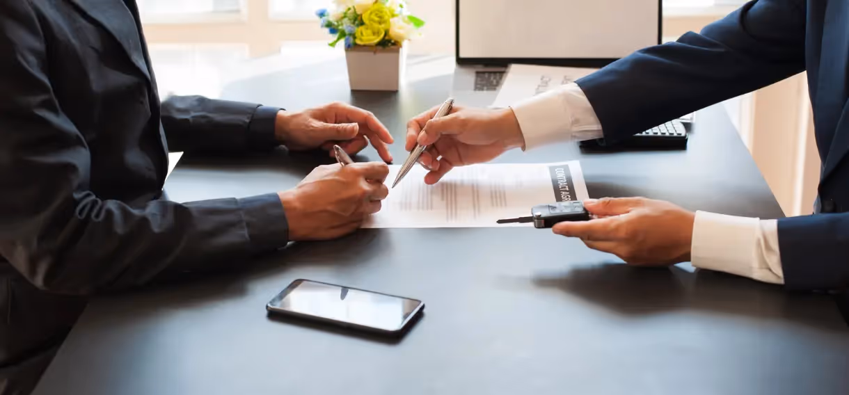 Two individuals in formal attire signing a contract at a desk with a phone and car key nearby.