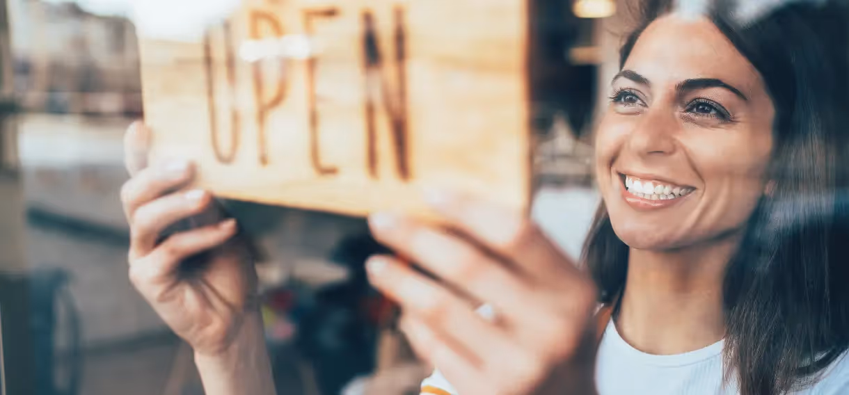 Smiling woman hanging a wooden sandblasted business sign with "OPEN" engraved on it inside a storefront window.