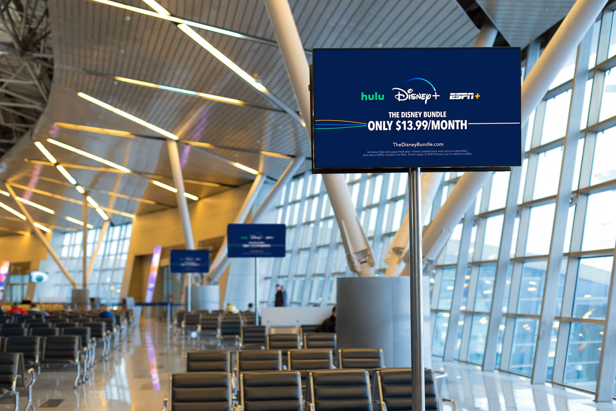 Digital DOOH advertising screen displaying Disney+ and Hulu ads in airport terminal seating area.