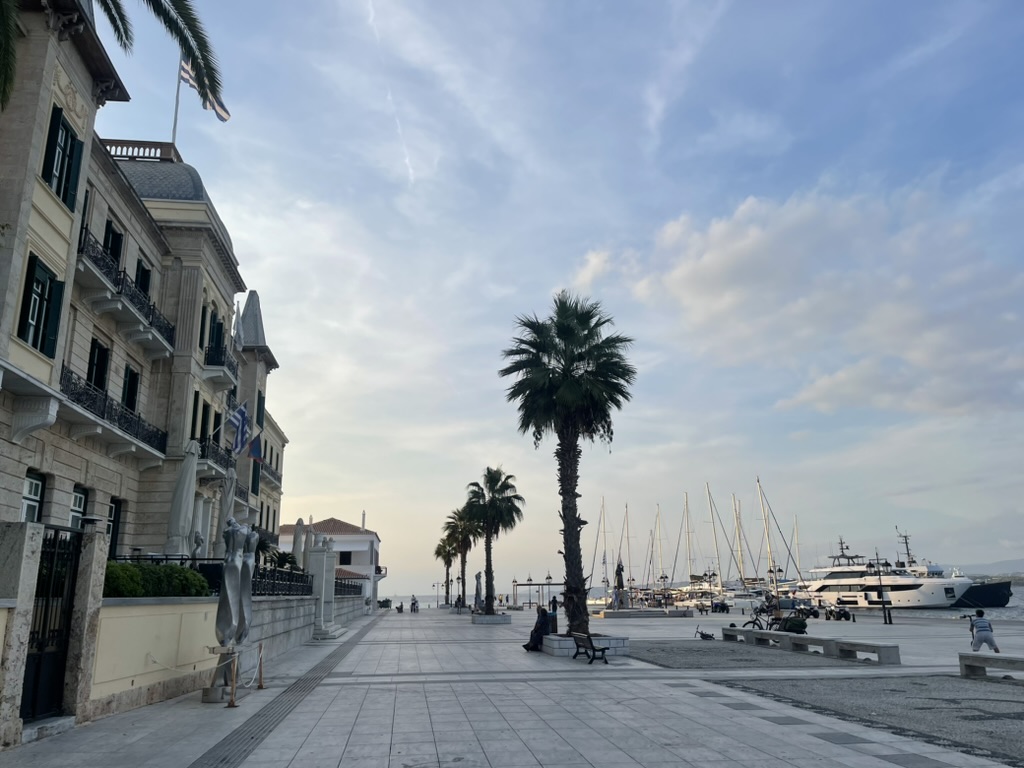 Een kadestraat met palmieren, banken en jachten aangemeerd in de haven onder een partly bewolkte blauwe lucht.