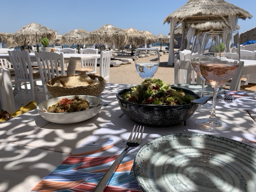 Tafel op het strand met mand brood, salade, glas rosé en water, omgeven door witte stoelen en rieten parasols.