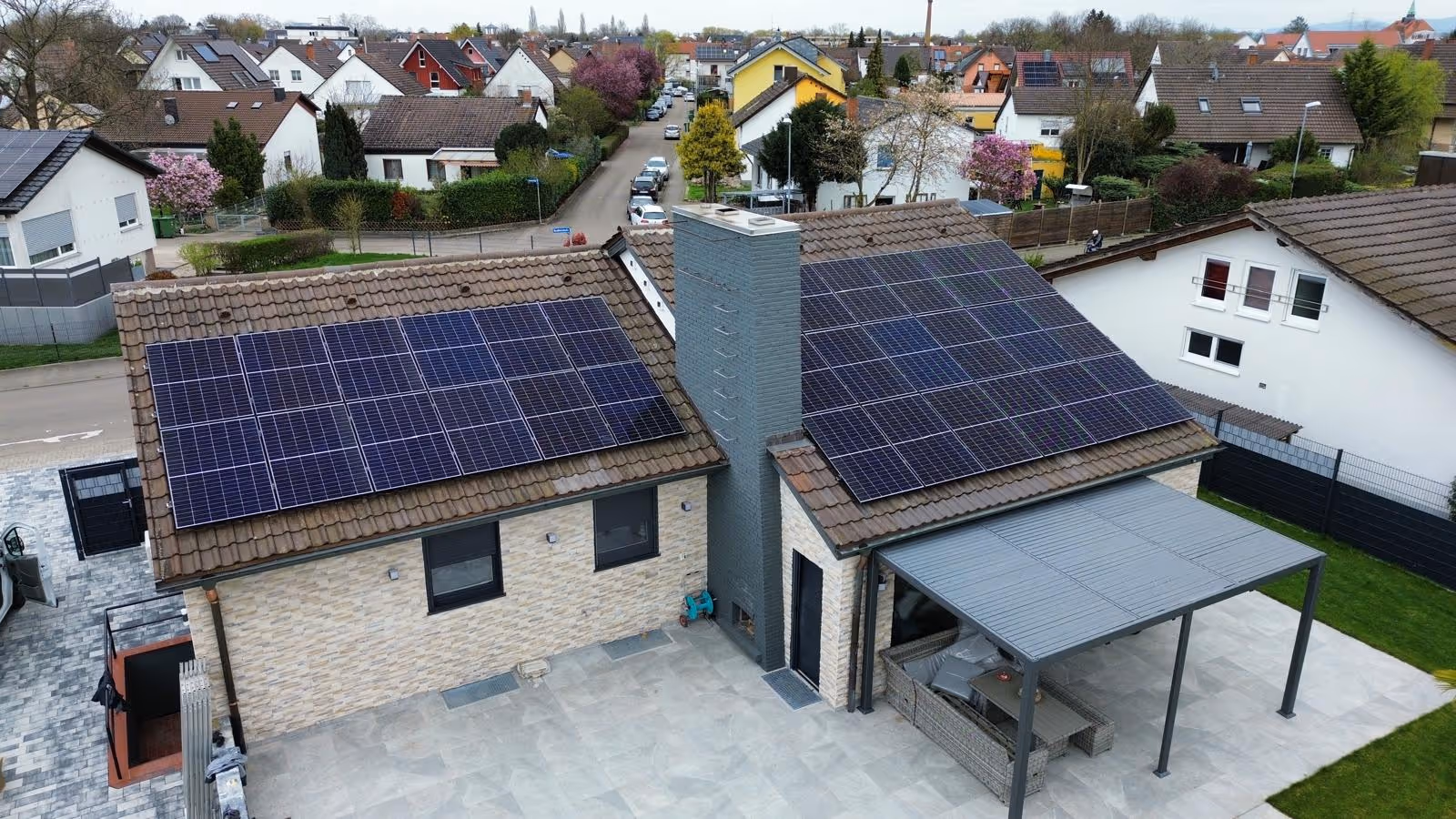 Zwei aneinandergebaute Häuser mit braunen Ziegeldächern und Solarpaneelen auf den Dächern, davor eine überdachte Terrasse mit Gartenmöbeln.