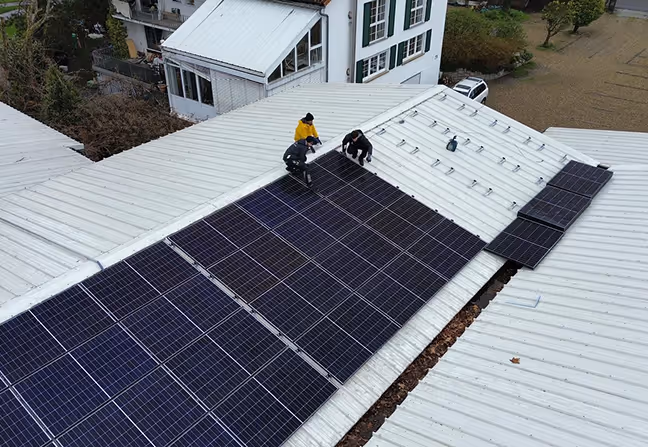 Drei Personen installieren Solarpaneele auf einem weißen Metallflachdach eines Gebäudes.