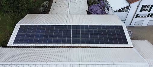 Dach mit Solarpaneelen, aufgenommen von oben, neben einem weißen Gebäude und einem Pflanzbereich mit lila Blumen.