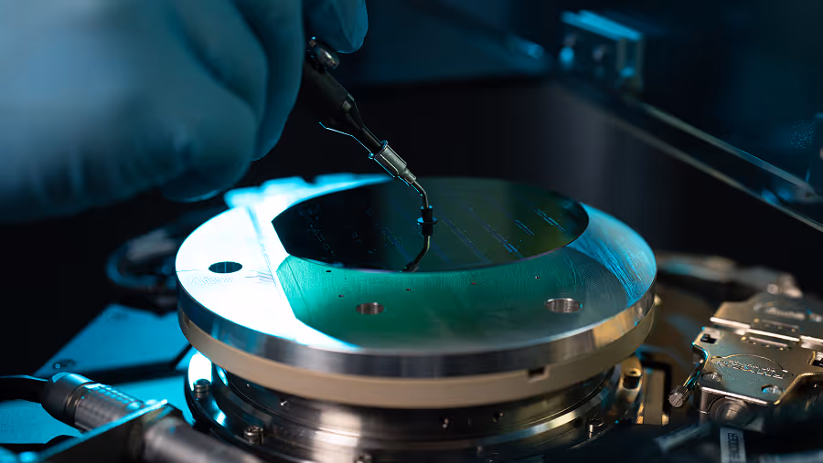 Gloved hand holding a precision probe near a silicon wafer on a high-tech testing device.