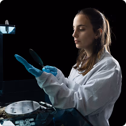 Scientist wearing a lab coat and blue gloves examining a silicon wafer in a dark laboratory.