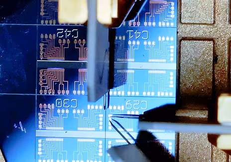 Close-up of electronic testing probes contacting a blue circuit board with visible microchip traces and labeled components.