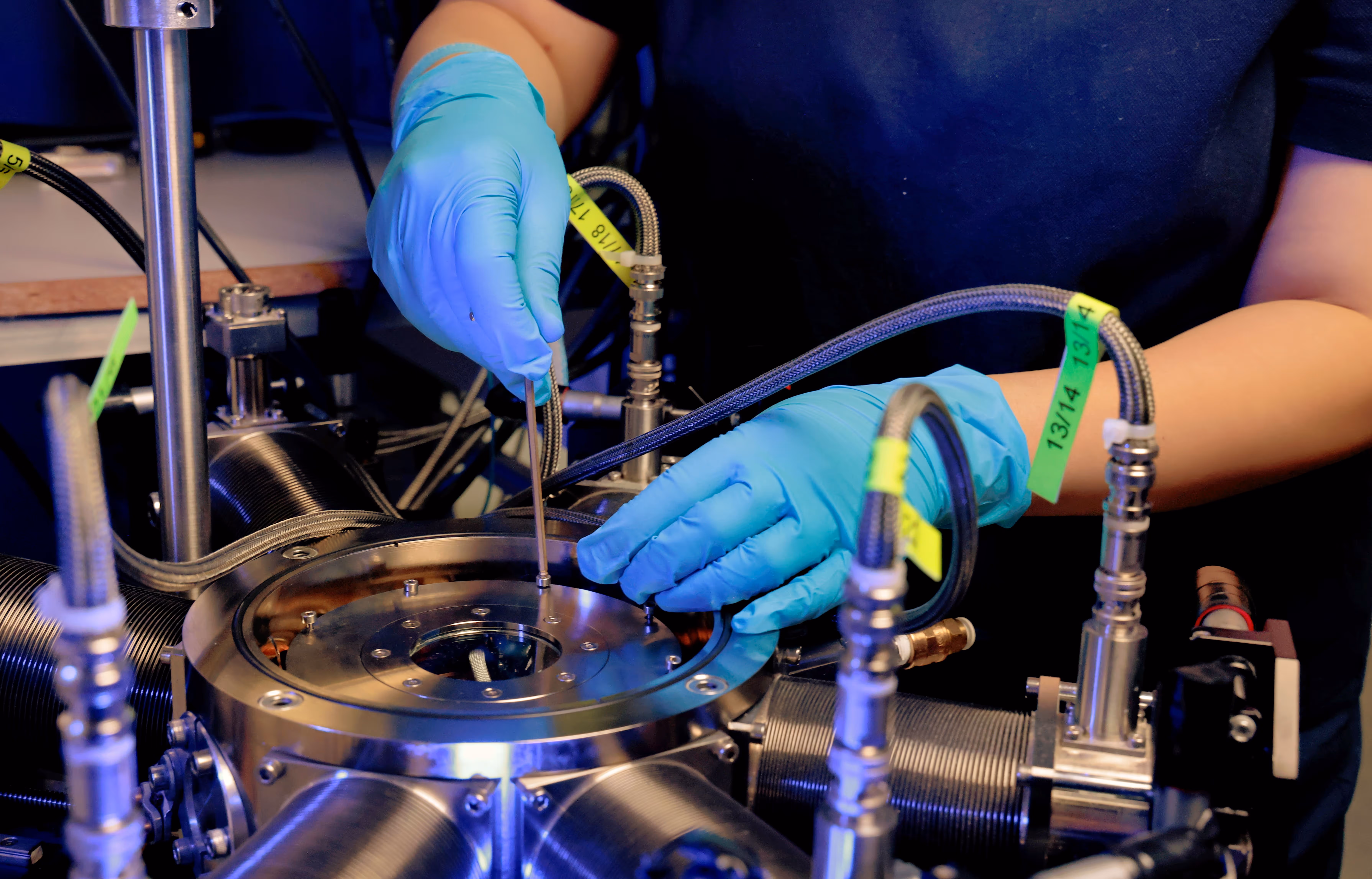 Person wearing blue gloves using a tool to adjust a valve or fitting on a metallic machine with multiple hoses attached.