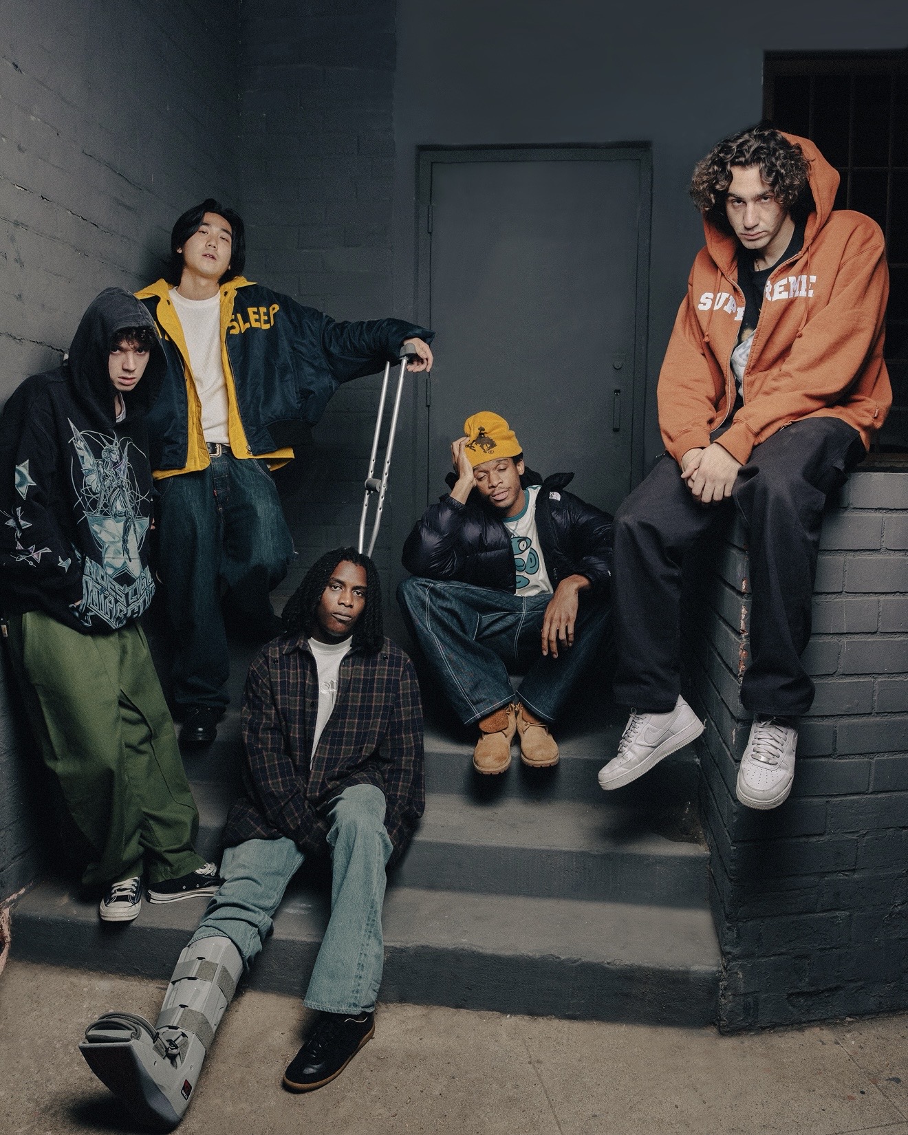 WHATMORE pose on concrete steps in a dim outdoor corridor, styled in hoodies, wide-leg jeans, and sneakers, with one member seated in front wearing a walking boot.