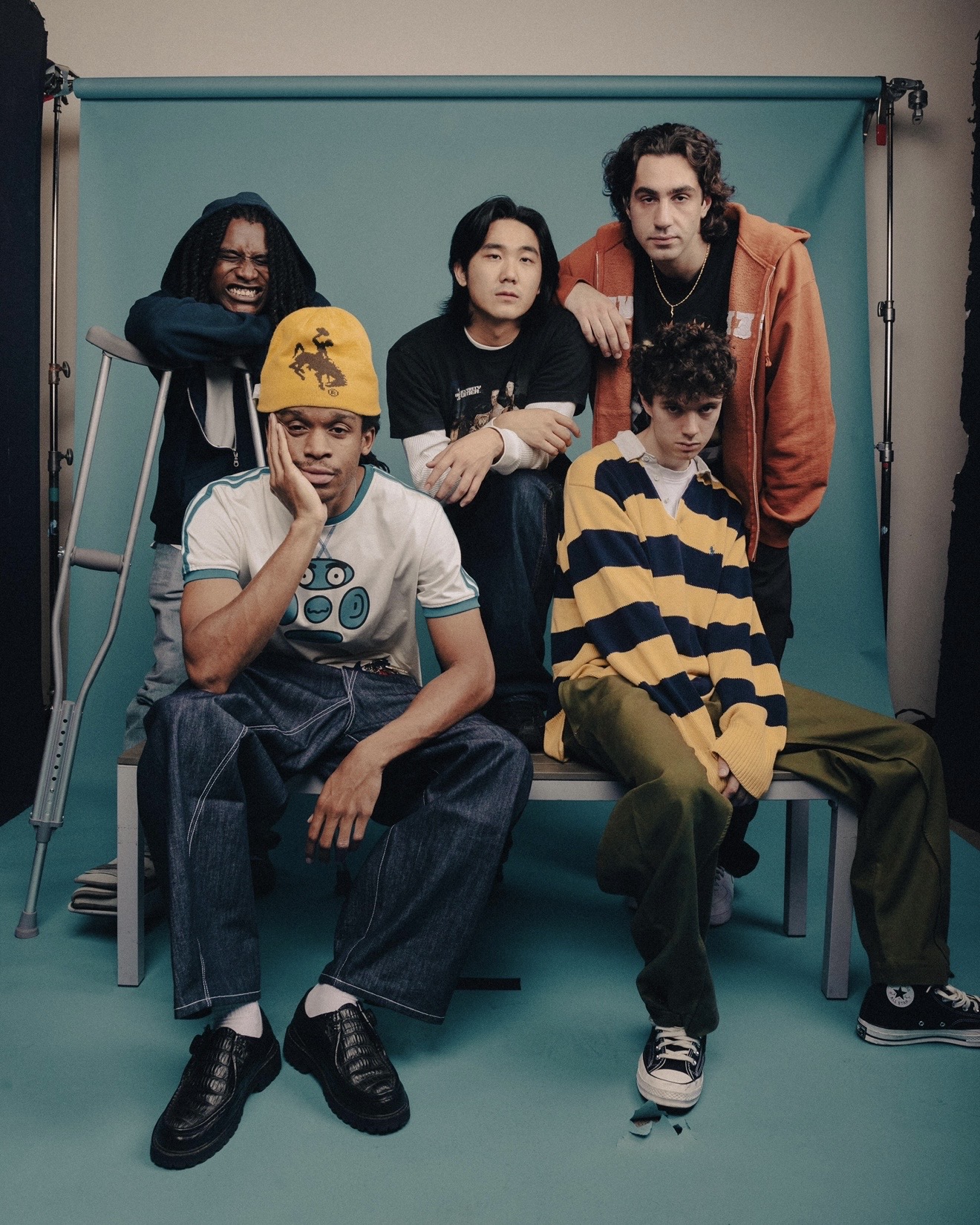 Wide studio portrait of WHATMORE seated and standing around a bench with a teal paper backdrop and light stands visible, styled in streetwear and sneakers.