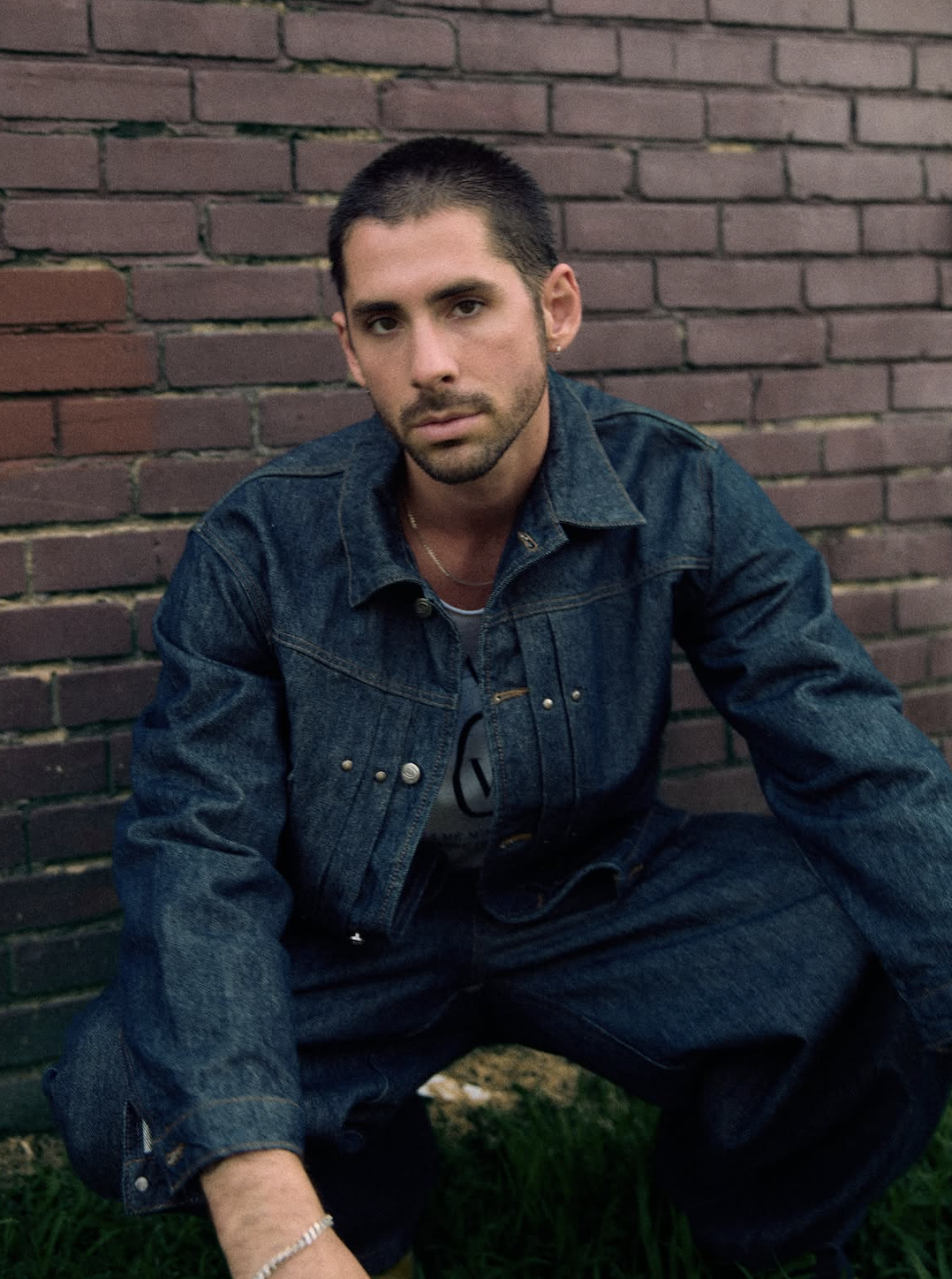 Mack Keane crouches against a brick wall in a full denim jacket and jeans, looking directly at the camera.