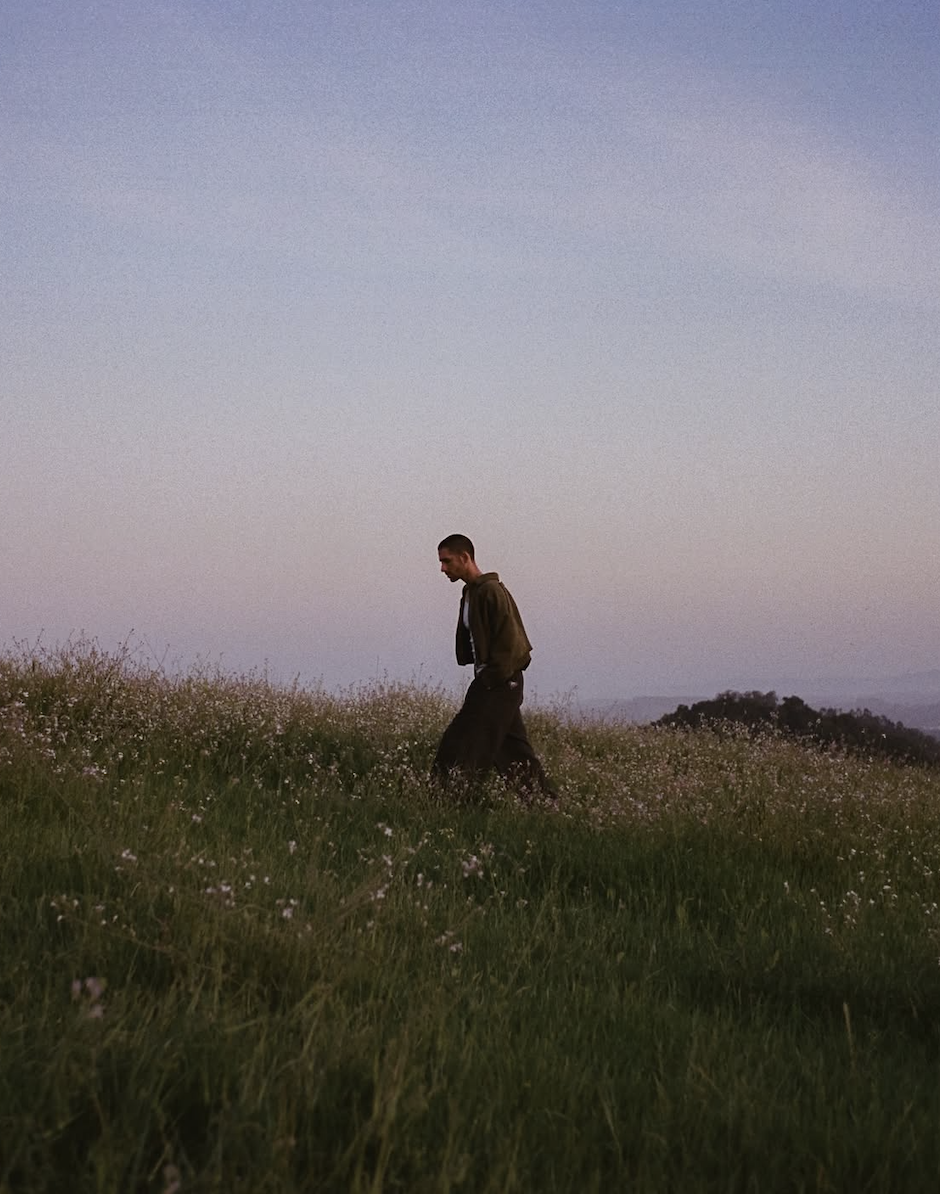 Mack Keane walks through a grassy hillside of wildflowers at dusk, hands in pockets, for his Wide Eyed interview with Medicine Box.