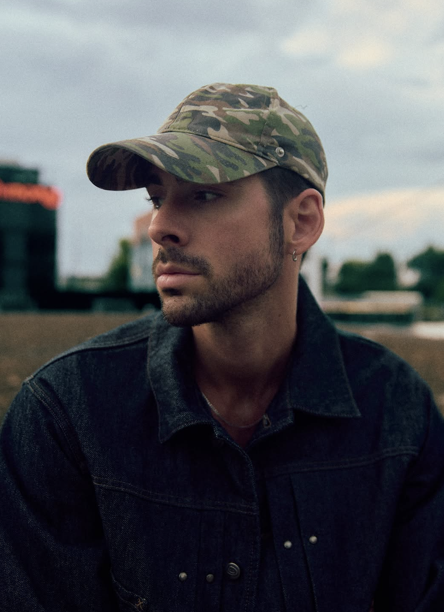 Close-up portrait of Mack Keane in a camouflage cap and dark denim jacket, looking to the side against a cloudy city skyline.