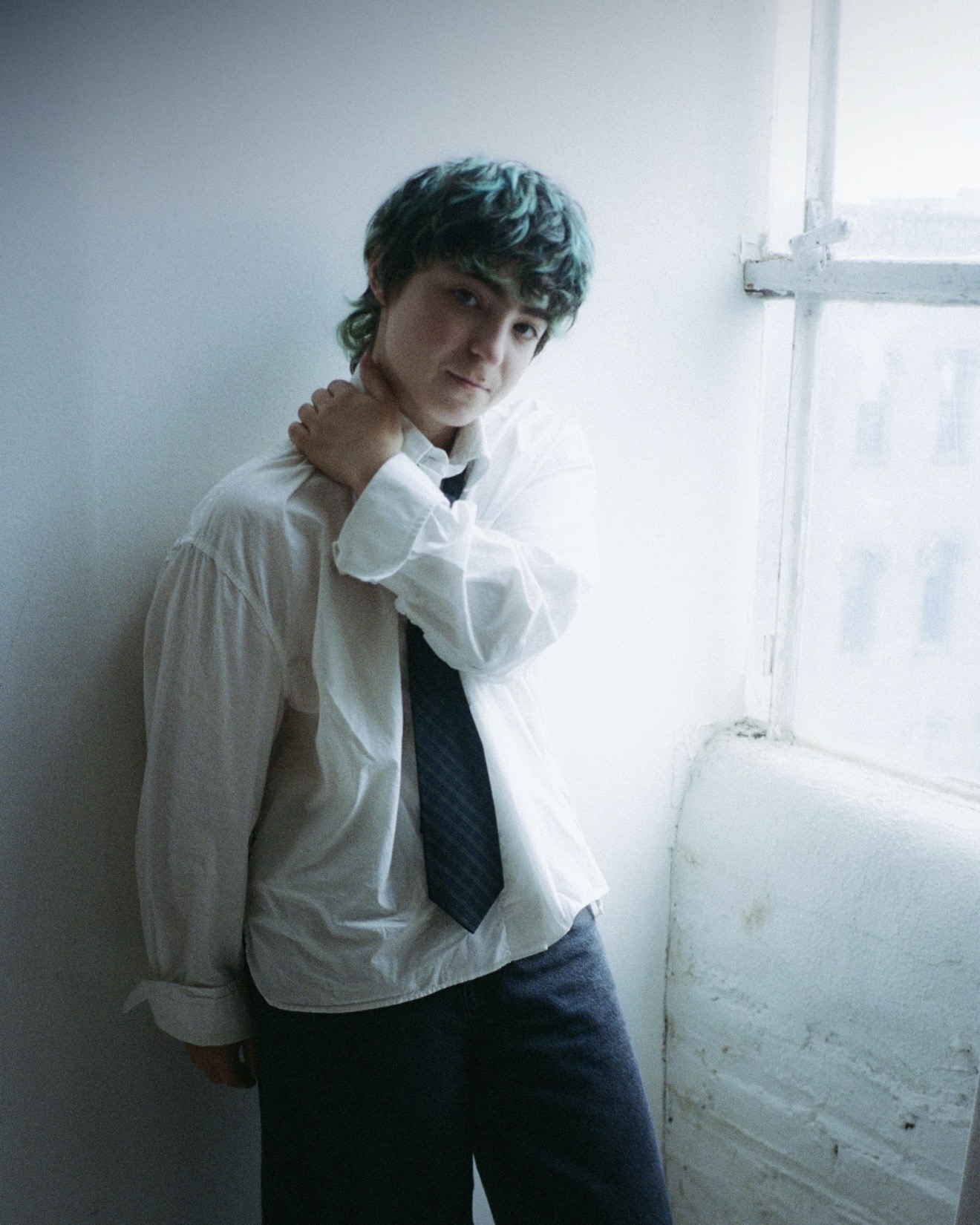 Claud leaning against a white wall by a window in an oversized white shirt and loose navy tie, hand on neck, Me and the Music interview and photoshoot, Medicine Box 2026