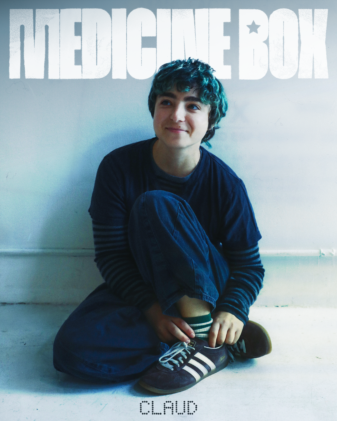 Claud sitting on the floor against a white wall in a navy t-shirt and striped sleeves, smiling off camera, Me and the Music album interview, Medicine Box 2026