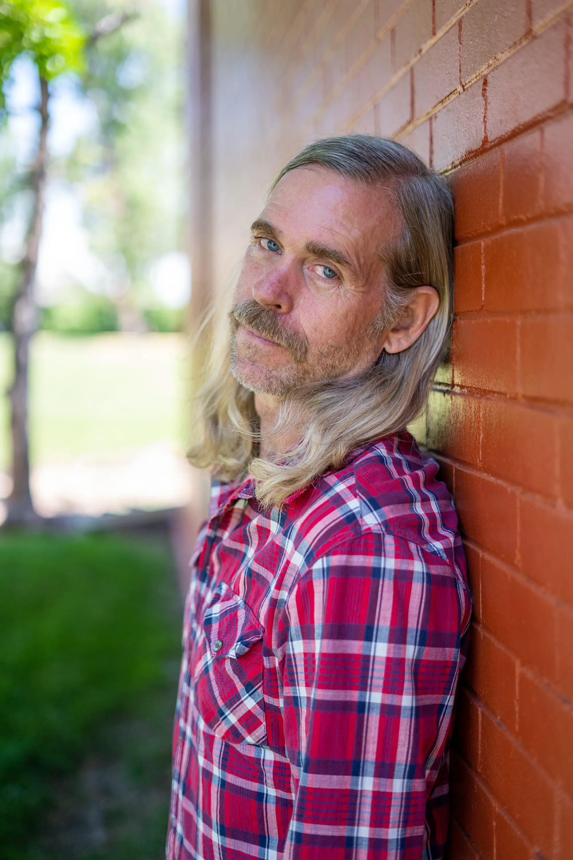 Man leaning against a brick wall with a red plaid shirt looking at the camera