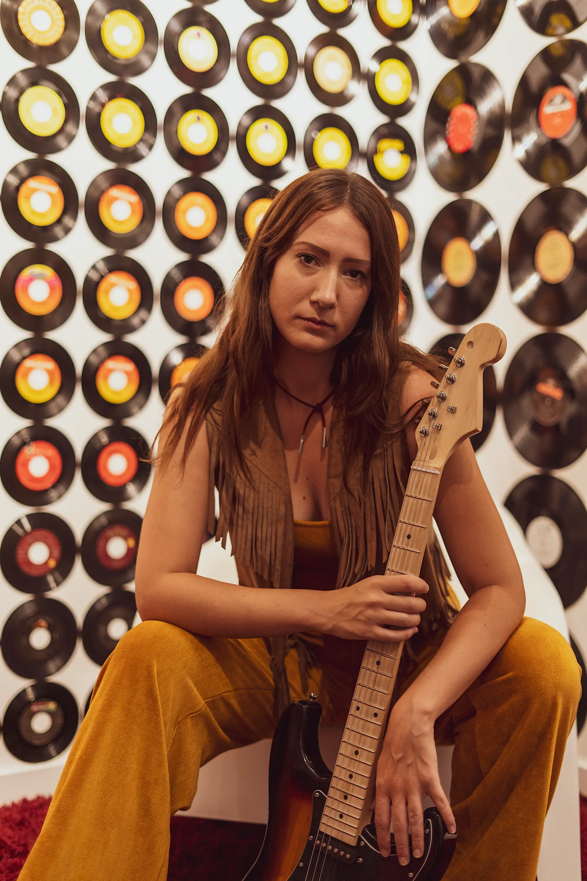 Woman in 1960's attire holding a guitar and looking at the camera with a wall of records behind her