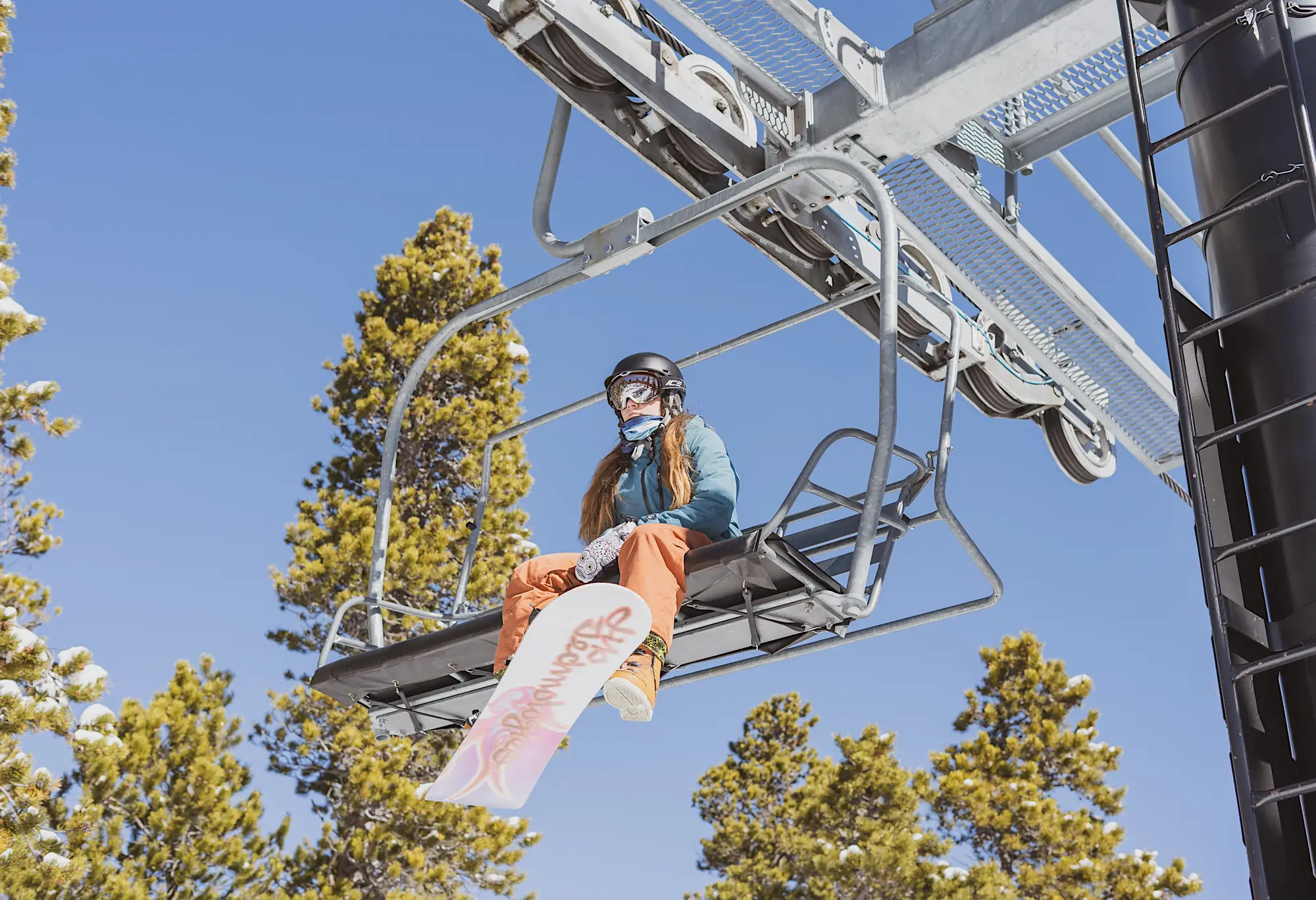 Woman sitting on a ski lift with a snowboard against green trees and a blue sky