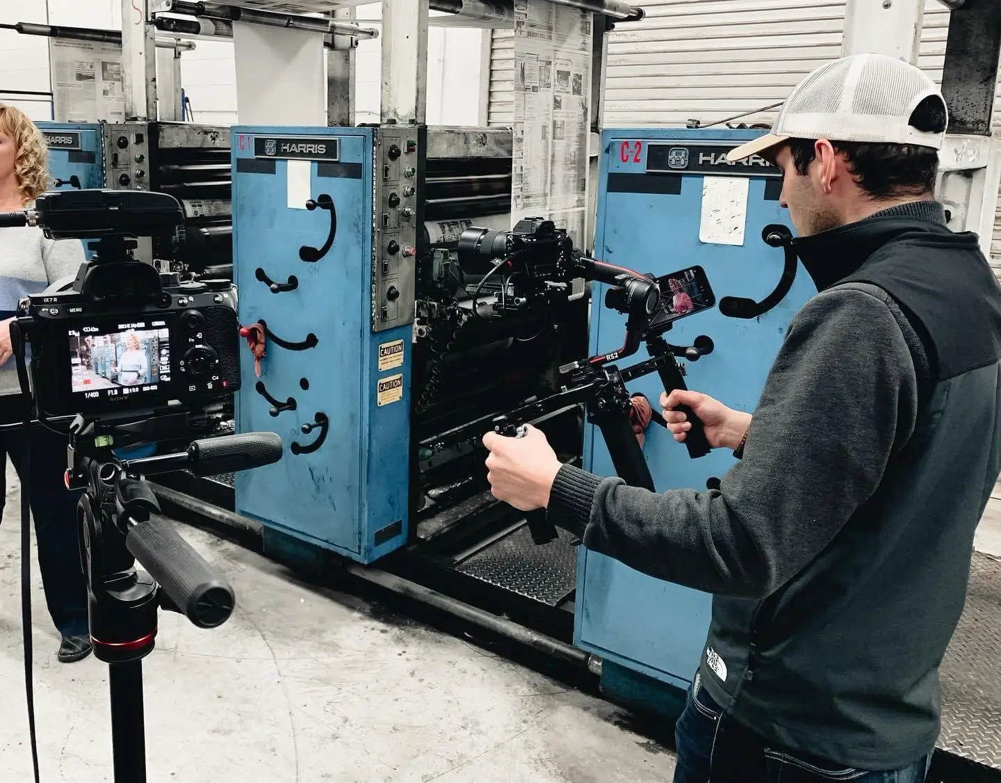 Filmmaker in dark vest and white cap operating a camera stabilizer to film a woman standing by industrial blue printing machinery.